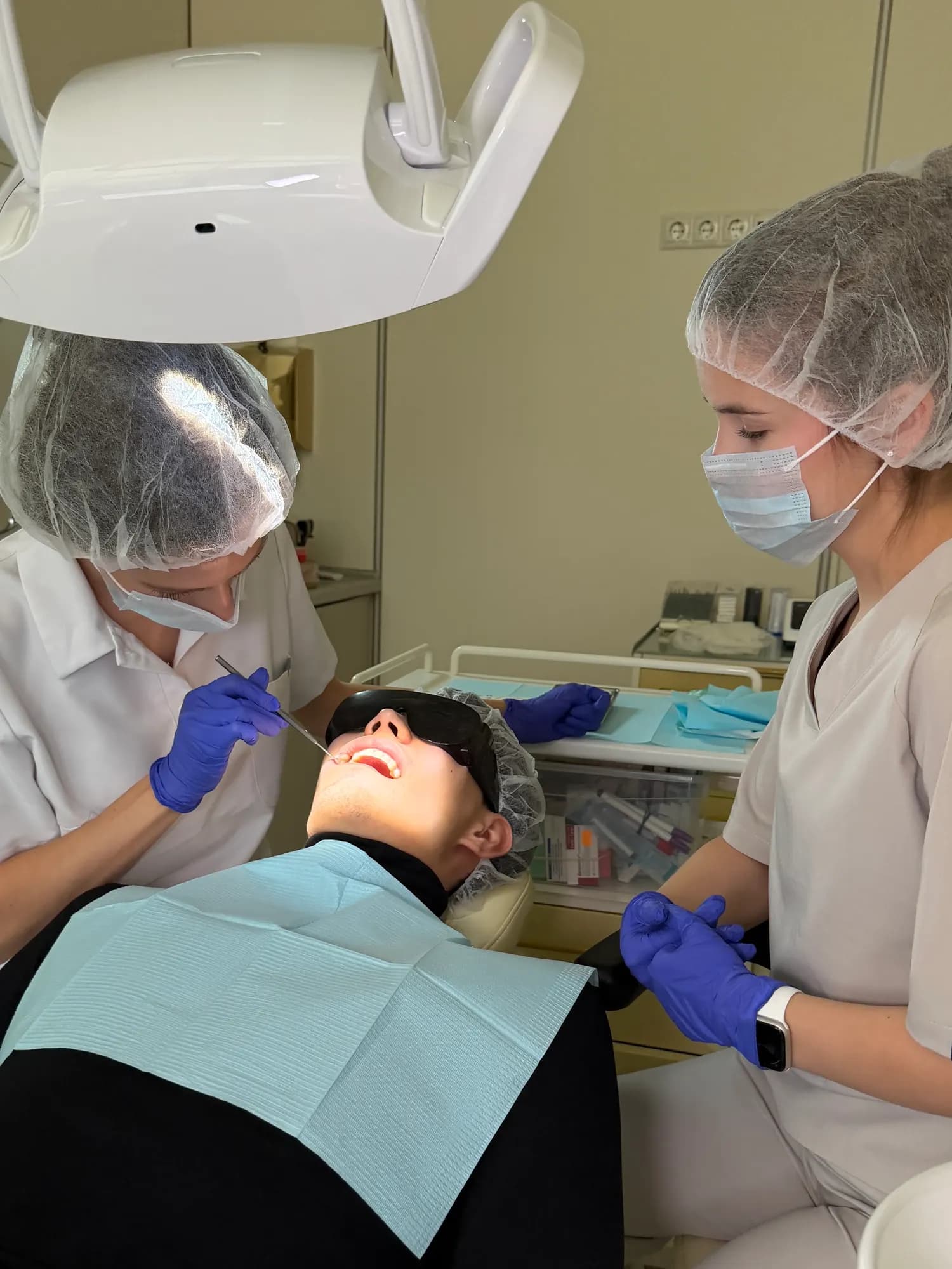 A dentist examines a patient's teeth with a dental mirror while an assistant stands by